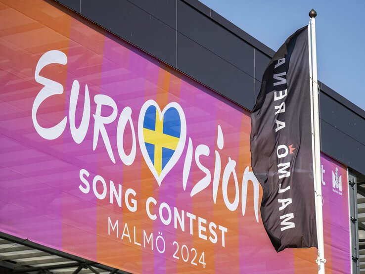 Banner des Eurovision Song Contest 2024 in Malmö mit Logo und schwedischer Flagge an einem Gebäude.